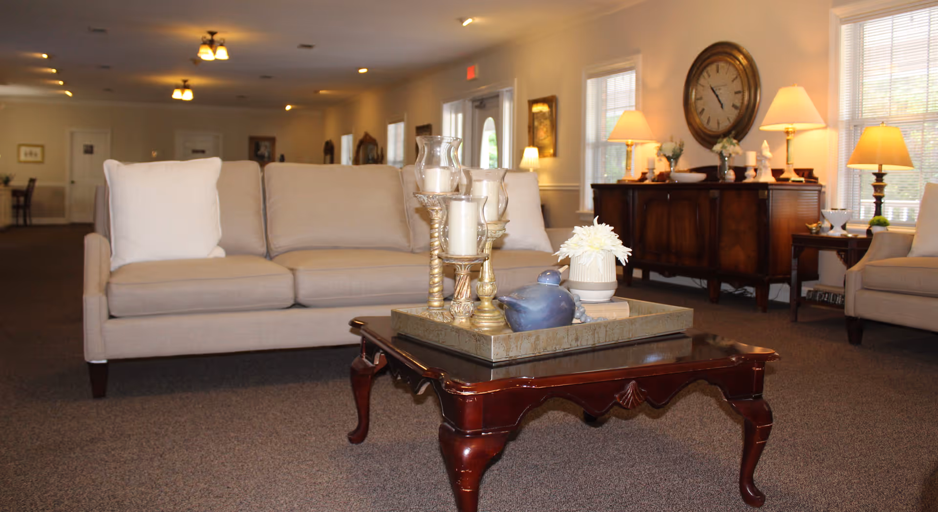 A cozy living room area in a retirement home featuring a beige sofa with white pillows, a wooden coffee table with decorative candles and a small vase with flowers, and a wooden sideboard with lamps and a large round clock on the wall. The room is warmly lit with multiple lamps and ceiling lights, and windows with blinds allow natural light to enter.