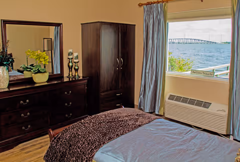 A bedroom with a bed covered in a blue and brown blanket, a dark wooden dresser with a mirror, decorative plants, and candles. There is a matching dark wooden wardrobe next to a window with blue curtains. Outside the window, a body of water and a bridge are visible.