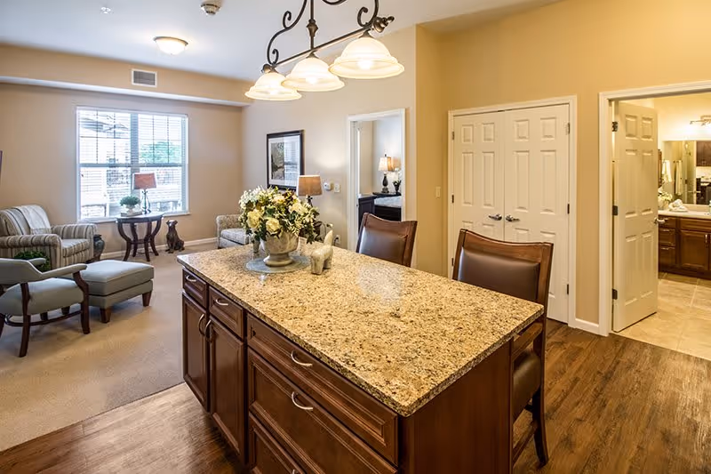 Interior view of a senior living facility showing a kitchen island with a granite countertop and two chairs. In the background, there is a living area with two armchairs, a small table with a lamp, and a large window letting in natural light. To the right, there are two open doorways leading to other rooms, one of which appears to be a bathroom.