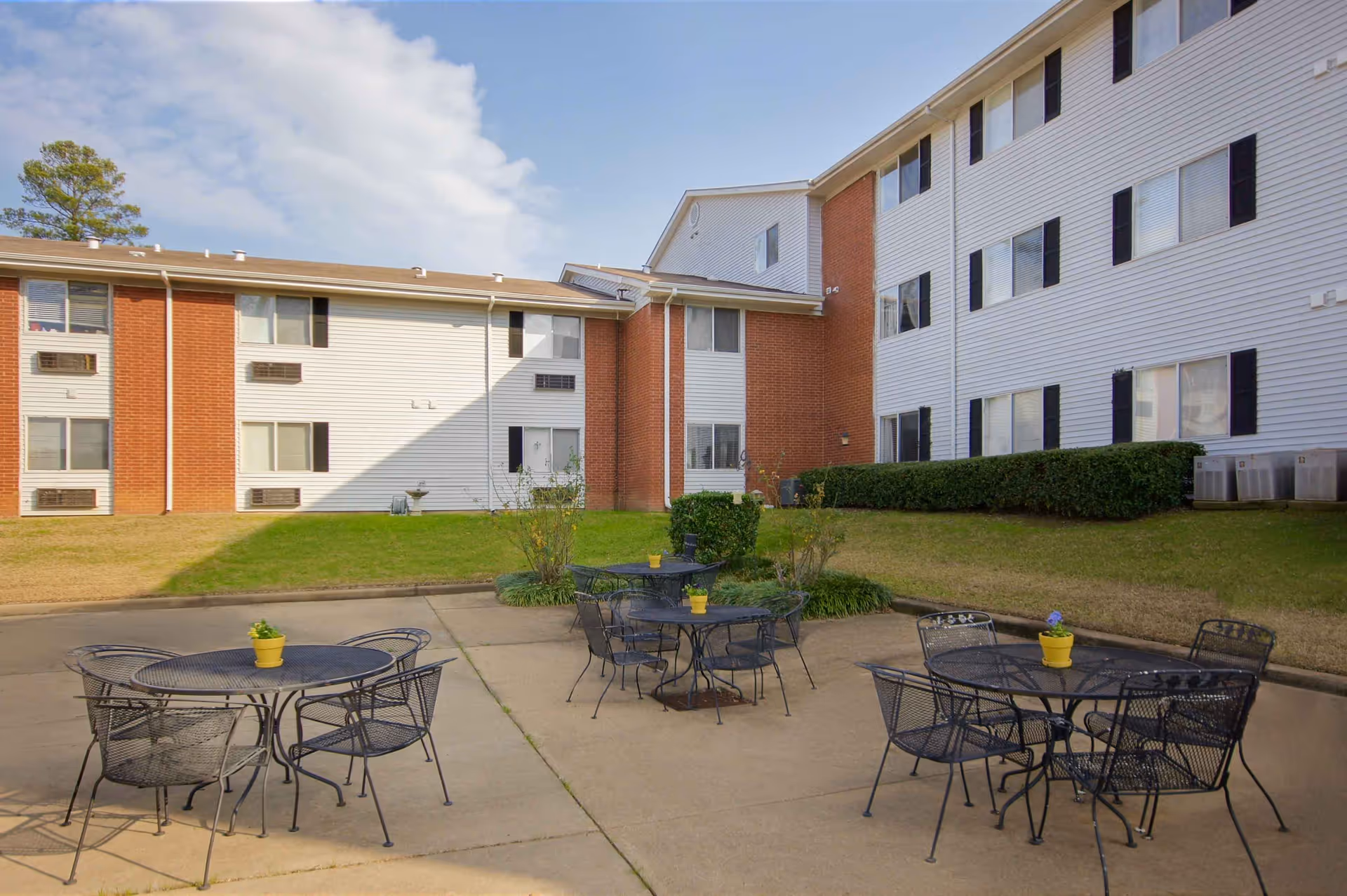 Outdoor patio area with several black metal tables and chairs, each table decorated with a small yellow flower pot. The patio is surrounded by a grassy area and a multi-story building with white siding and red brick accents under a partly cloudy sky.