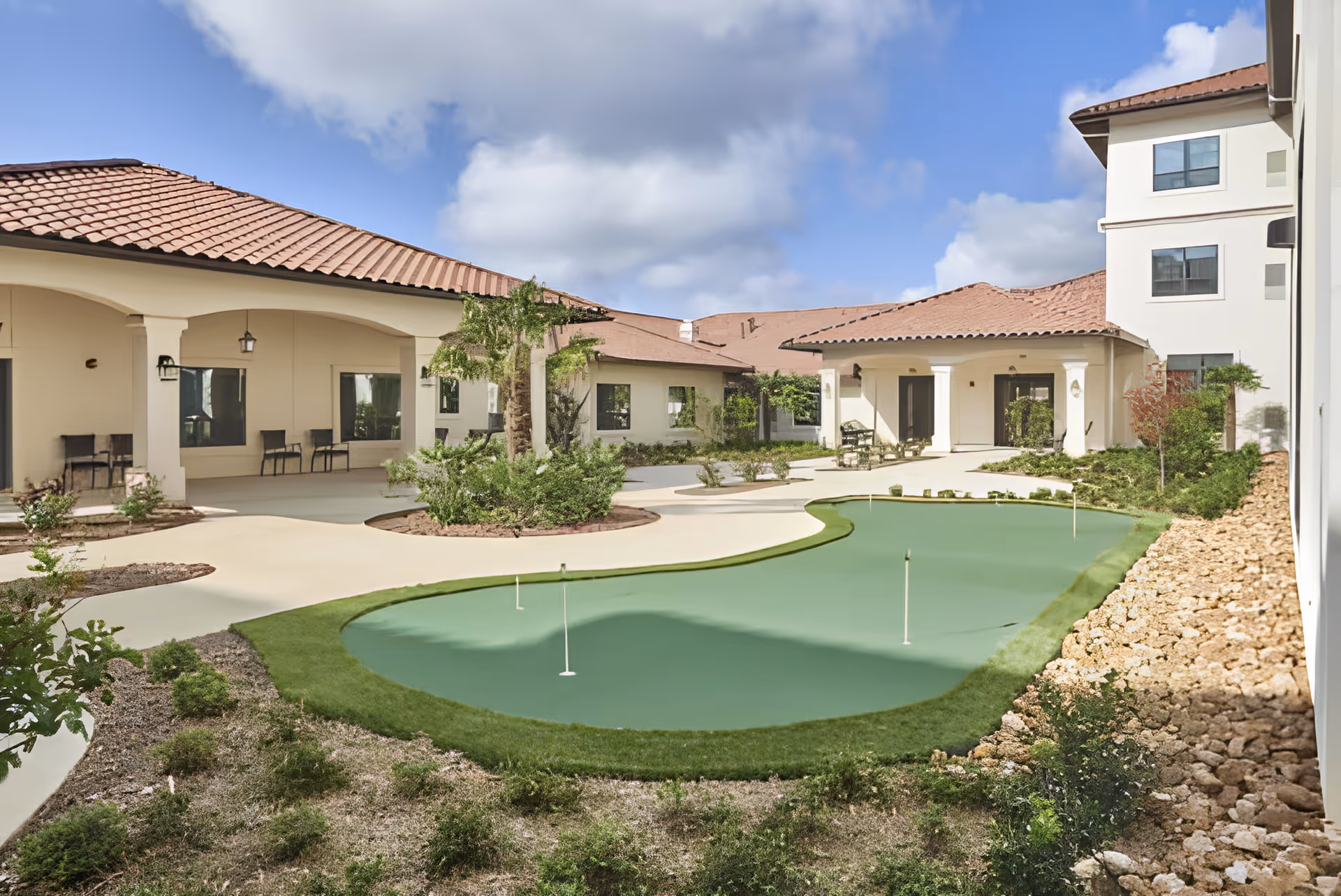 Courtyard with a small putting green surrounded by covered walkways and Mediterranean-style assisted living buildings under a partly cloudy sky.