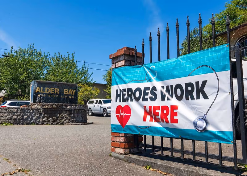 Entrance gate of Alder Bay Assisted Living facility with a banner that reads 'HEROES WORK HERE' featuring a red heart with a heartbeat line and a stethoscope graphic. A stone sign with the facility name is visible in the background along with parked vehicles and trees under a clear blue sky.