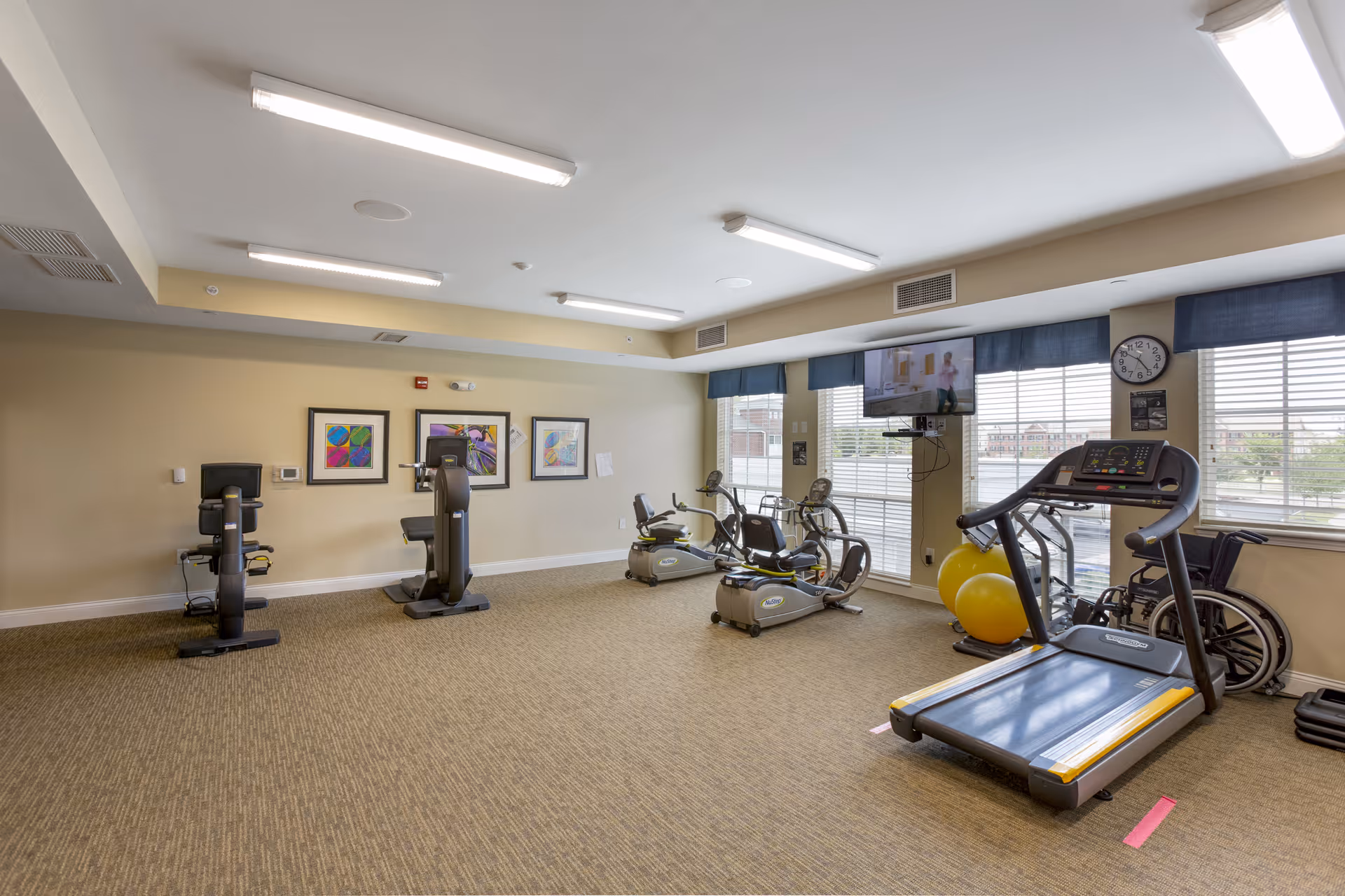 A fitness room in a senior living facility with exercise equipment including a treadmill, stationary bikes, and exercise balls. The room has beige walls, carpeted floor, three framed colorful artworks on the wall, large windows with blue valances, and a wall-mounted TV. A wheelchair is positioned near the treadmill.