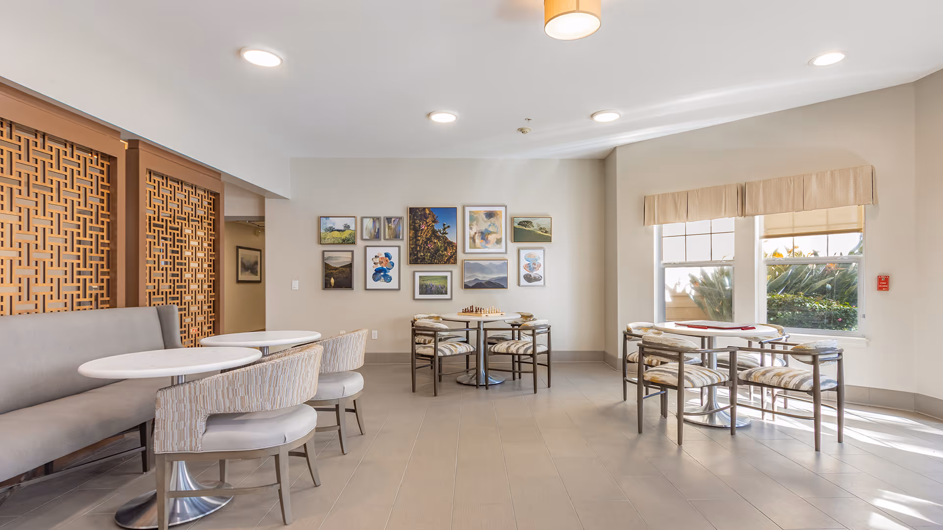 Well-lit communal dining area with round tables, upholstered chairs, wall art, and large windows letting in natural light.