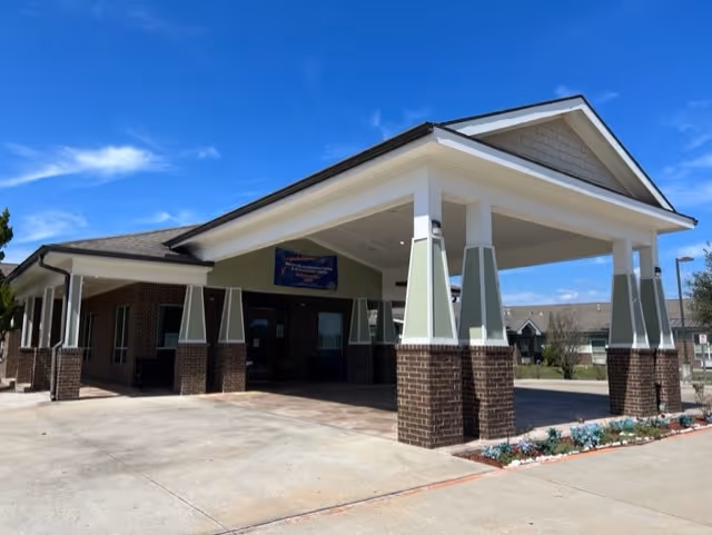 Exterior view of Arbor Hills Rehabilitation and Healthcare Center showing the main entrance with a covered drop-off area supported by brick and light green pillars under a clear blue sky.
