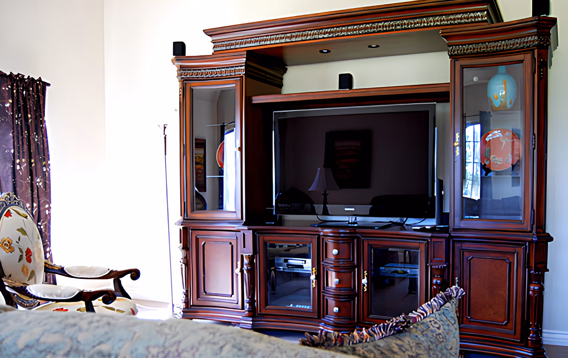 A living room area featuring a large wooden entertainment center with a flat-screen TV in the center. The entertainment center has glass cabinets displaying decorative vases. To the left, there is a floral upholstered armchair and a floor lamp. A patterned cushion is visible in the foreground.