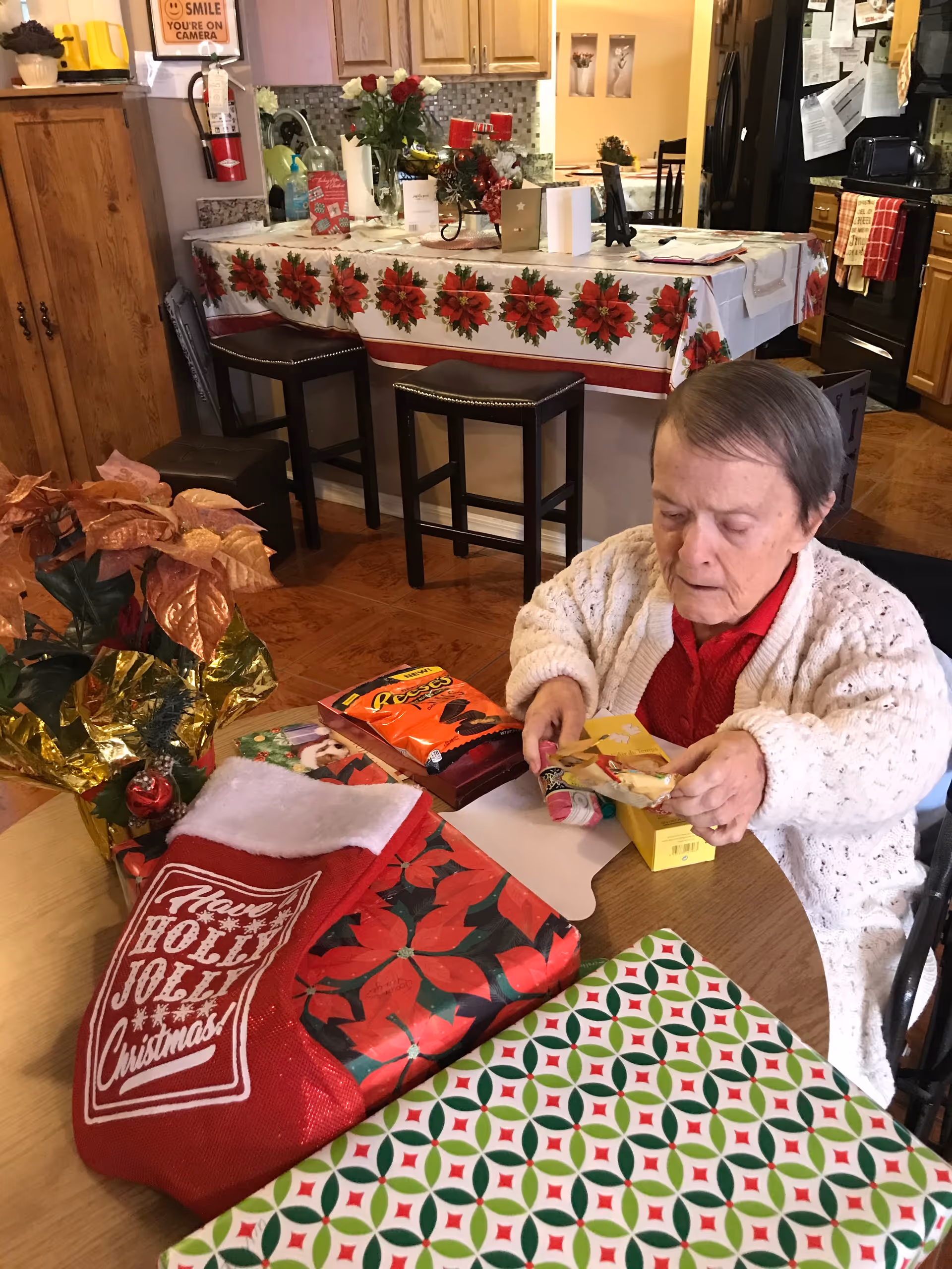 An elderly woman wearing a white knitted cardigan and red shirt is sitting at a round table in a kitchen or dining area. The table is decorated with Christmas-themed items including a red stocking with 'Have a Holly Jolly Christmas!' written on it, wrapped gifts, and holiday decorations. Behind her, there is a kitchen counter with a poinsettia-themed tablecloth, stools, and kitchen appliances visible in the background.