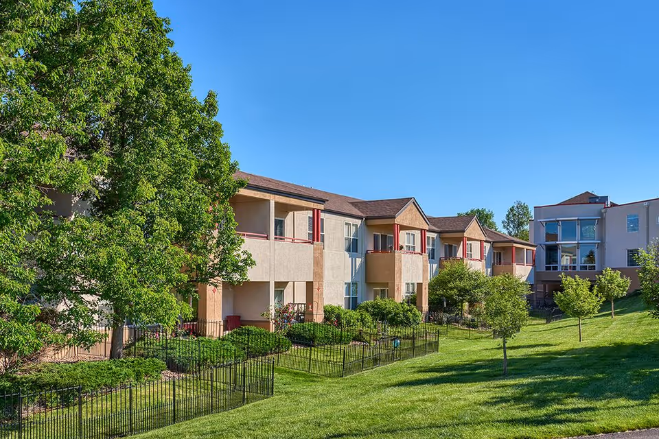 Exterior view of a two-story senior living building with balconies, landscaped lawns, and trees under a clear blue sky.