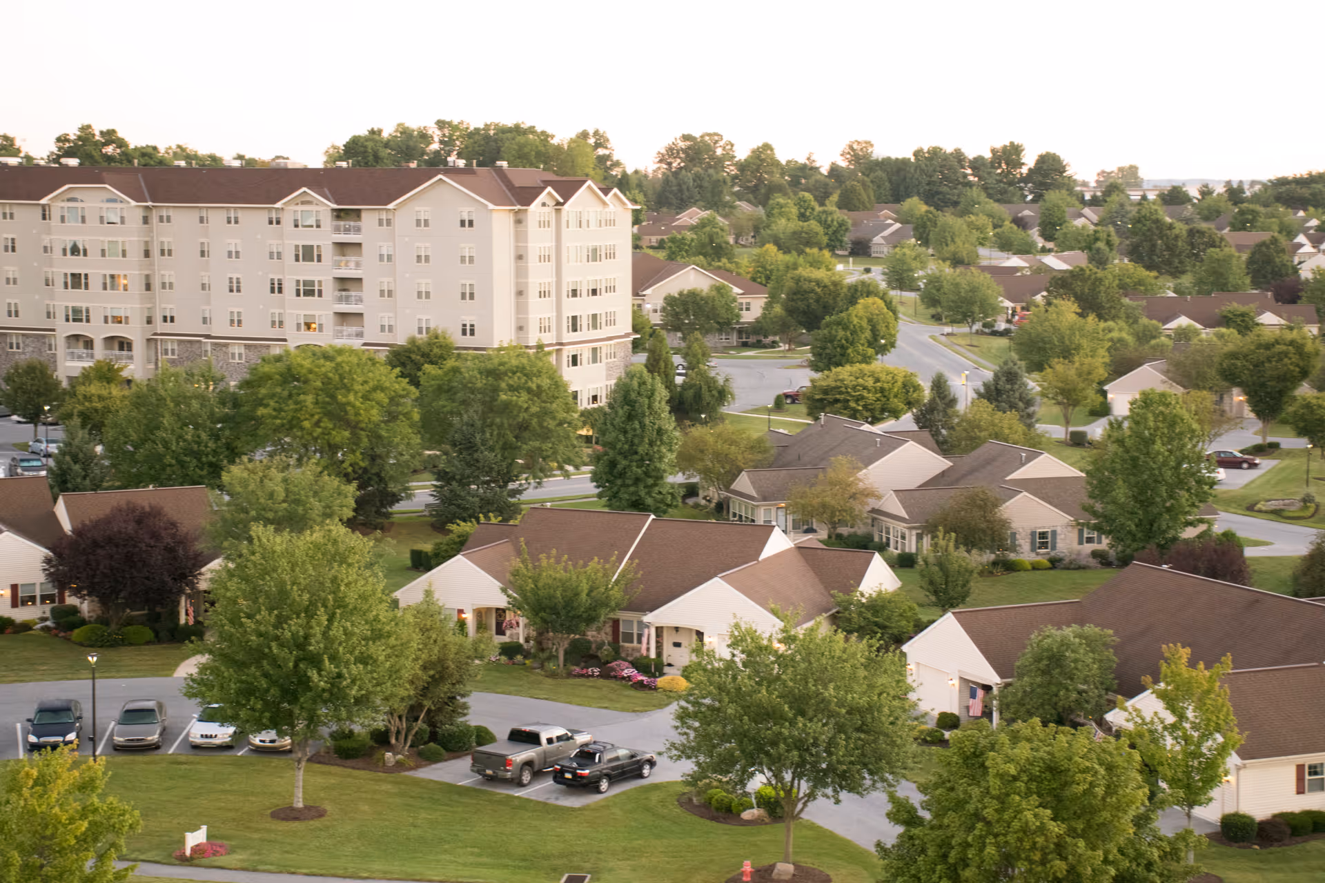 Aerial view of a senior living community with single-story cottages, a multi-story building, trees, and parked cars.