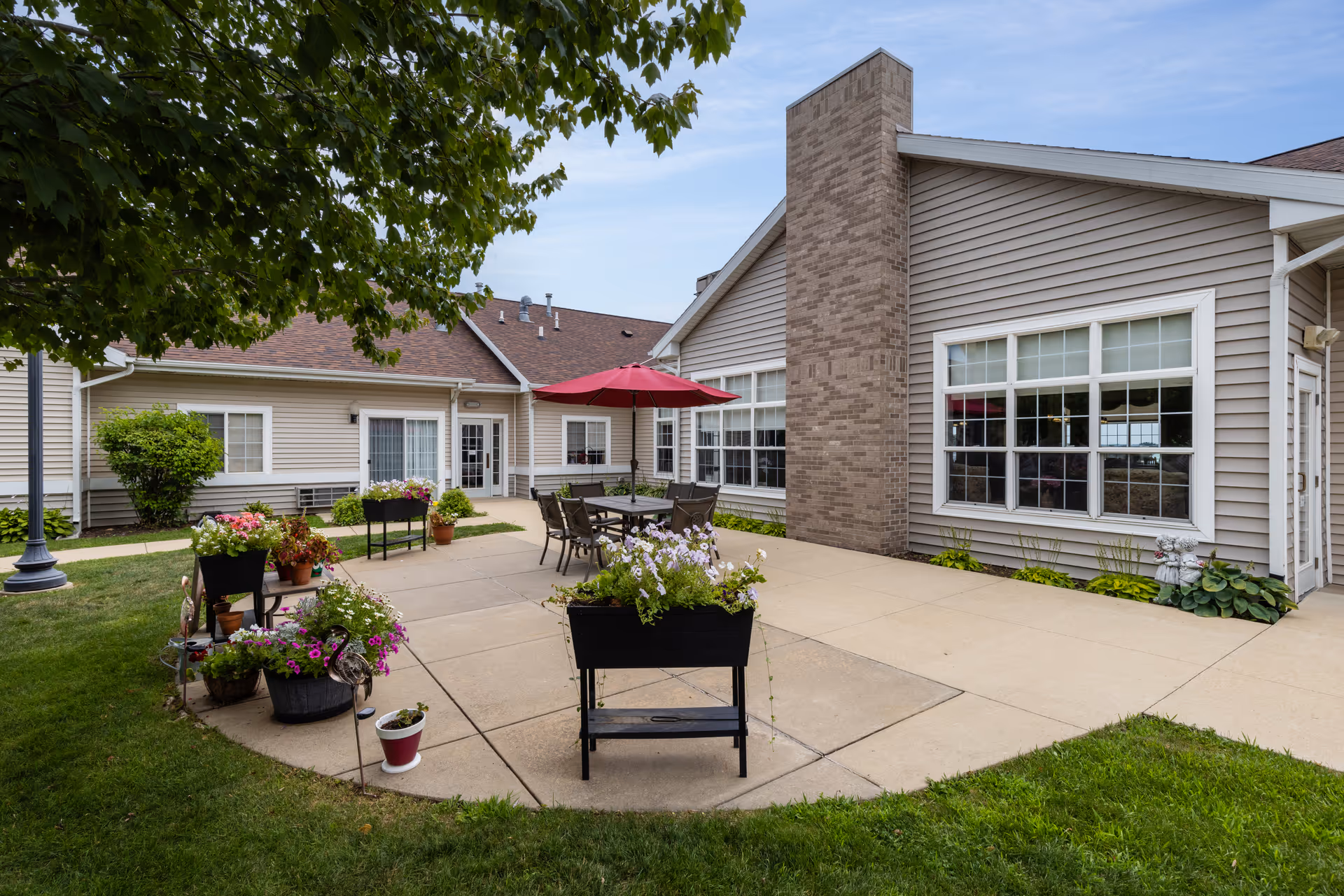 Outdoor patio area at Clover Ridge Place featuring a concrete seating area with a table, chairs, and a red umbrella. The patio is surrounded by green grass, potted flowers, and plants. The building exterior has beige siding, large windows, and a brick chimney. A tree and a lamp post are visible on the left side.