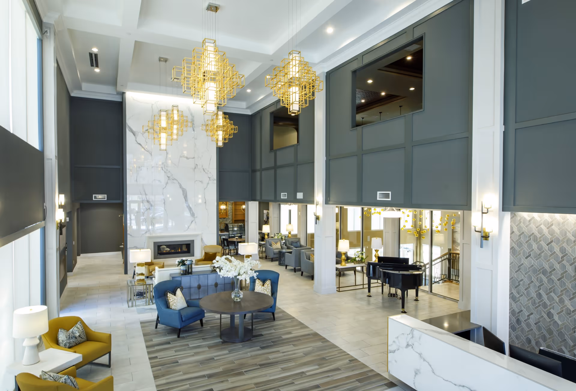 Spacious two-story assisted living lobby with seating areas, gold chandeliers, a marble fireplace, and a grand piano.