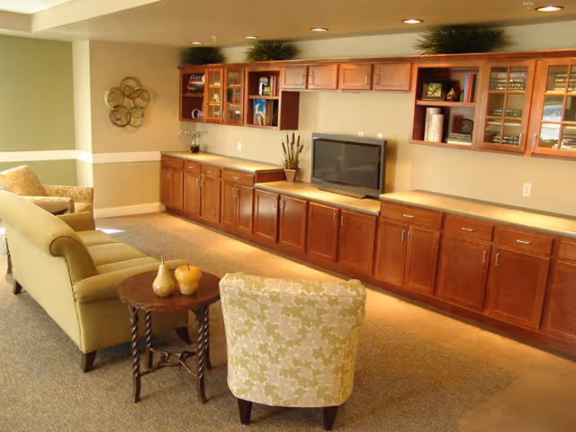 A cozy living room area with a green sofa, a patterned armchair, and a small wooden round table with decorative items. The room features a long wooden cabinet with multiple drawers and glass-front upper cabinets along one wall, with a flat-screen TV placed on the countertop. The walls are painted in a light beige color with white trim, and there is a decorative wall piece on the left side.