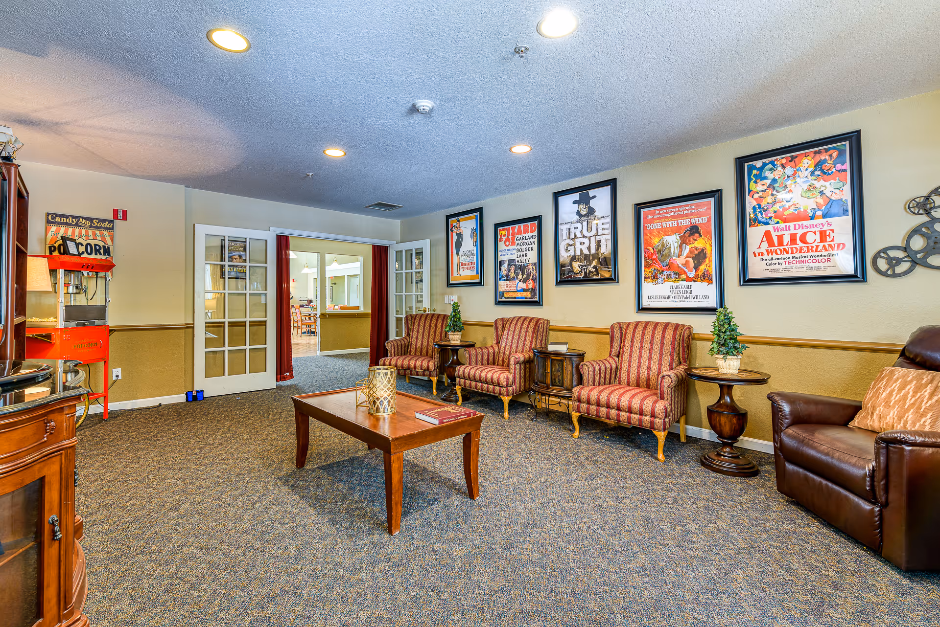 A cozy living room area with patterned armchairs, a leather recliner, a wooden coffee table with decorative items, and framed vintage movie posters on the wall. There is a popcorn machine in the corner and double glass doors leading to another room.
