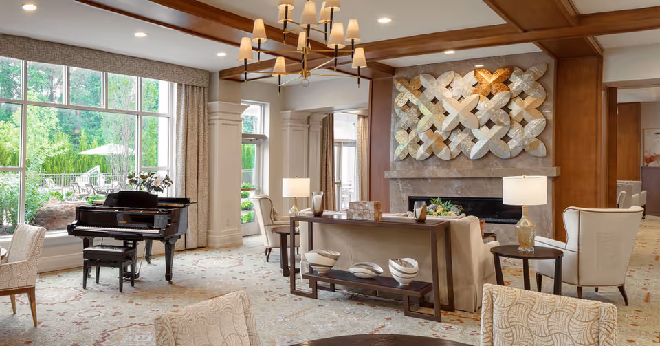 Bright, elegant senior living lounge with large windows, a grand piano, multiple seating areas, and a fireplace with decorative wall art.