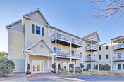 Three-story pale yellow senior apartment building with covered balconies, a main entrance, and a paved driveway under a clear blue sky.