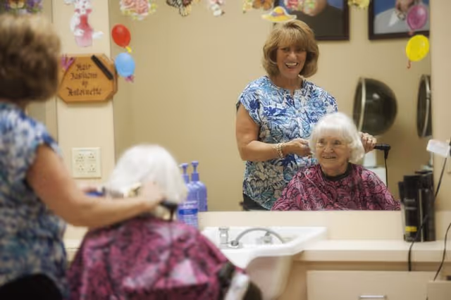 A smiling woman in a blue floral shirt is styling the white hair of an elderly woman seated in front of a mirror in a salon or grooming area. The elderly woman is wearing a purple cape. The room is decorated with colorful balloons and a sign on the wall.