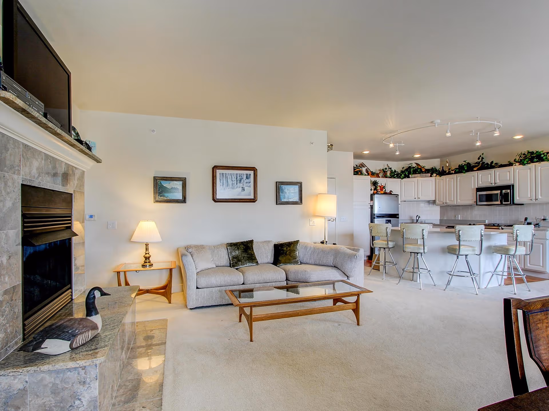A bright and spacious living room area with a light gray sectional sofa, glass coffee table, and two lamps on either side. The room features a fireplace with a decorative duck figurine on the hearth. In the background, there is an open kitchen with white cabinets, a refrigerator, microwave, and a breakfast bar with four cushioned bar stools. The ceiling has track lighting and there are framed pictures on the wall above the sofa.