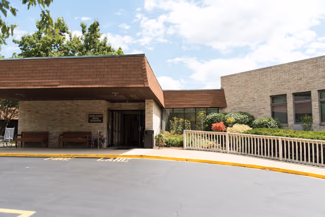 Covered entrance of a one-story brick senior living facility with benches, landscaped planters and a driveway drop-off.