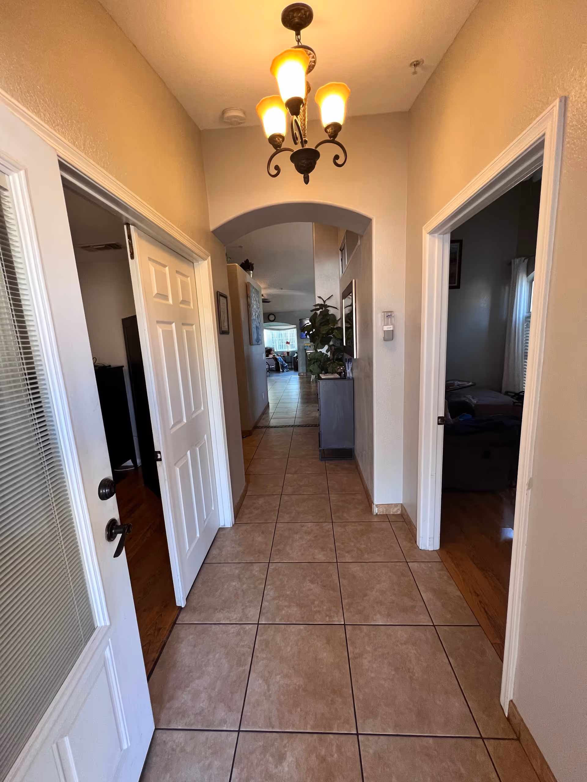 Tiled entry hallway with an arched doorway and chandelier leading into a living area, with doors to rooms on either side.