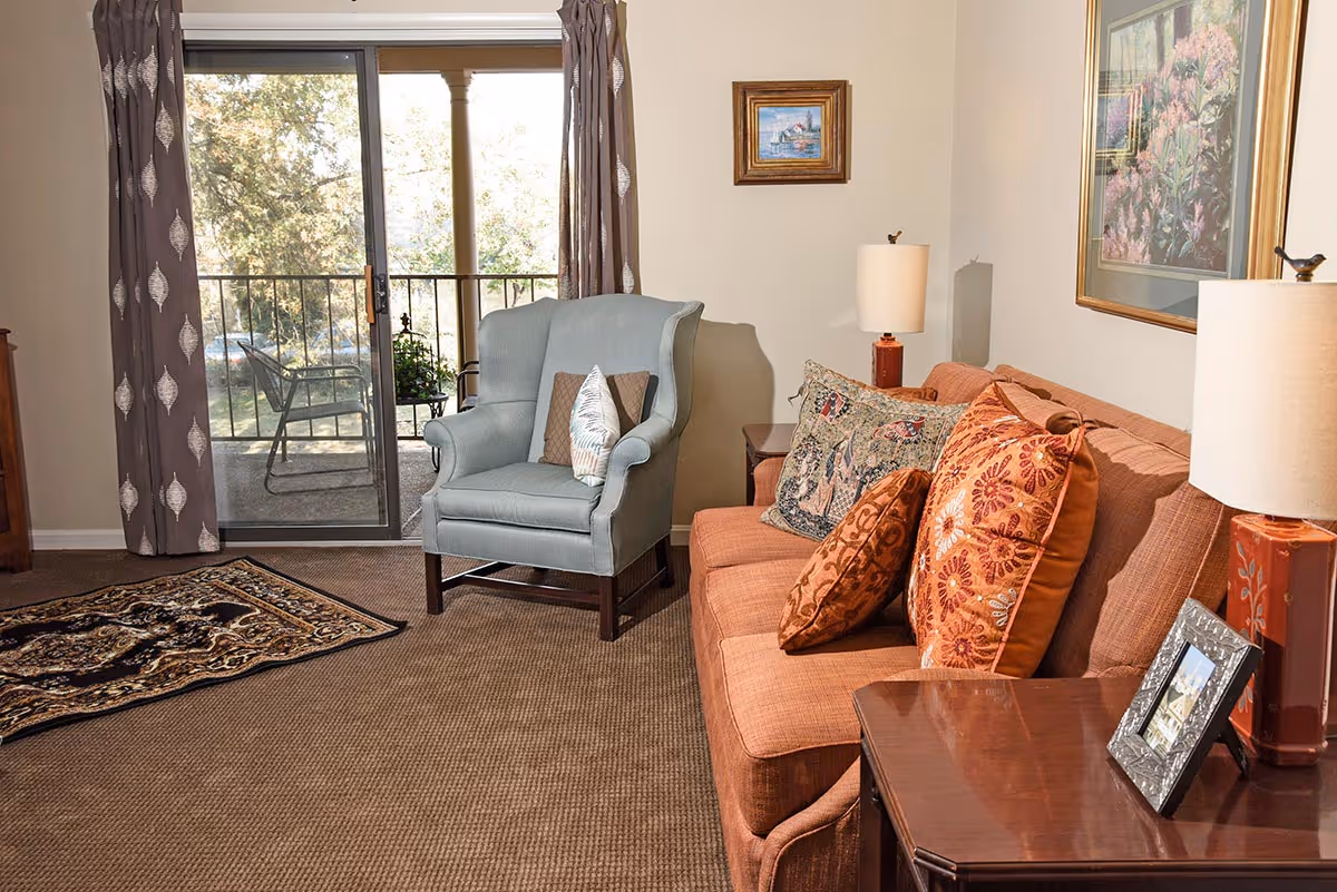 Cozy living room with an orange sofa and patterned pillows, a blue armchair, side tables with lamps, and a sliding glass door opening to a small balcony.