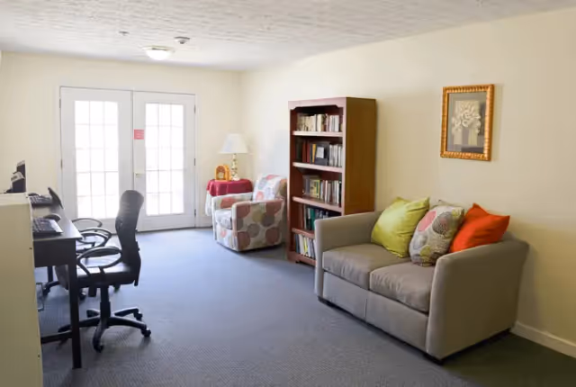 A cozy living room area with a gray sofa adorned with colorful pillows, a patterned armchair next to a small table with a lamp, a wooden bookshelf filled with books, and two office chairs at desks near a set of glass double doors letting in natural light.