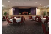 A large room with round tables covered with white tablecloths and surrounded by red cushioned chairs. At the far end of the room is a stage with a piano and some plants on either side. The room has a patterned carpet and recessed lighting in the ceiling.