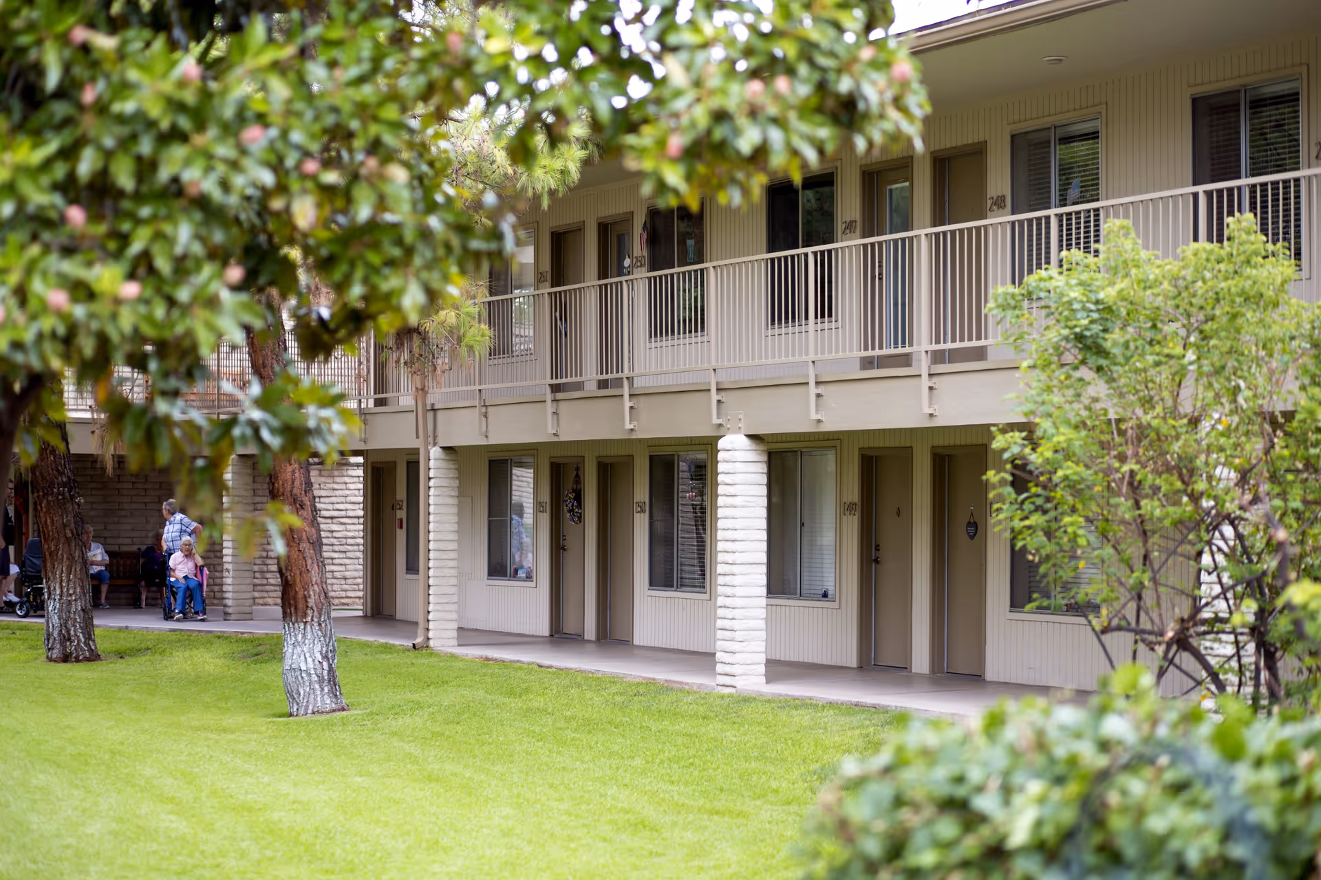 Exterior view of a two-story senior living facility building with a green lawn and trees in the foreground. Several doors and windows are visible on both floors, and a few elderly people are sitting and standing near the building under a covered area.