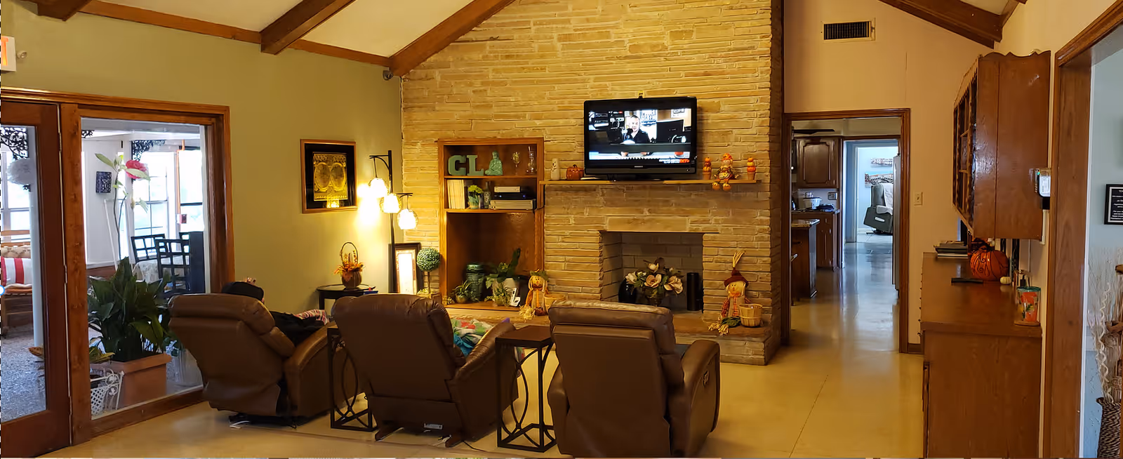 A cozy living room with three brown recliner chairs facing a stone fireplace with a mounted flat-screen TV above it. The room features wooden beams on the ceiling, a wooden cabinet on the right, and a doorway leading to another room. There are plants and decorative items around the fireplace and a glass door on the left leading to a sunroom with additional seating.