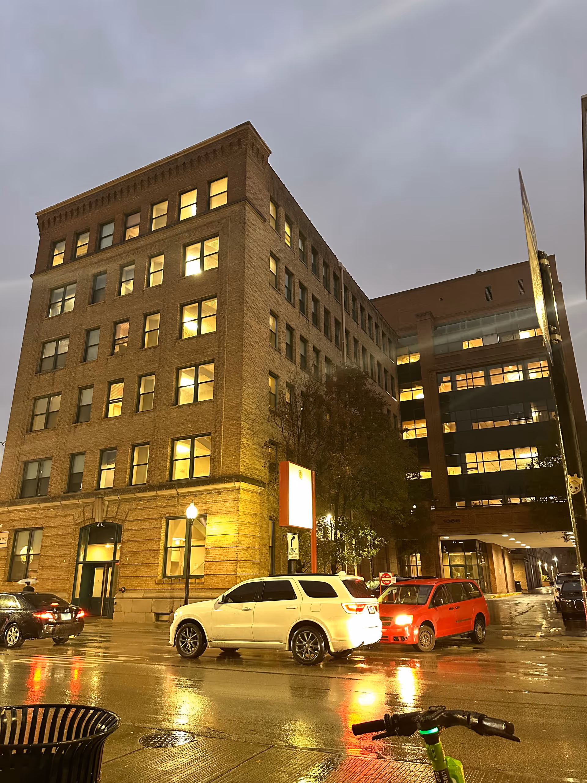 Multi-story brick building at night with illuminated windows, a wet street, and parked cars in front.