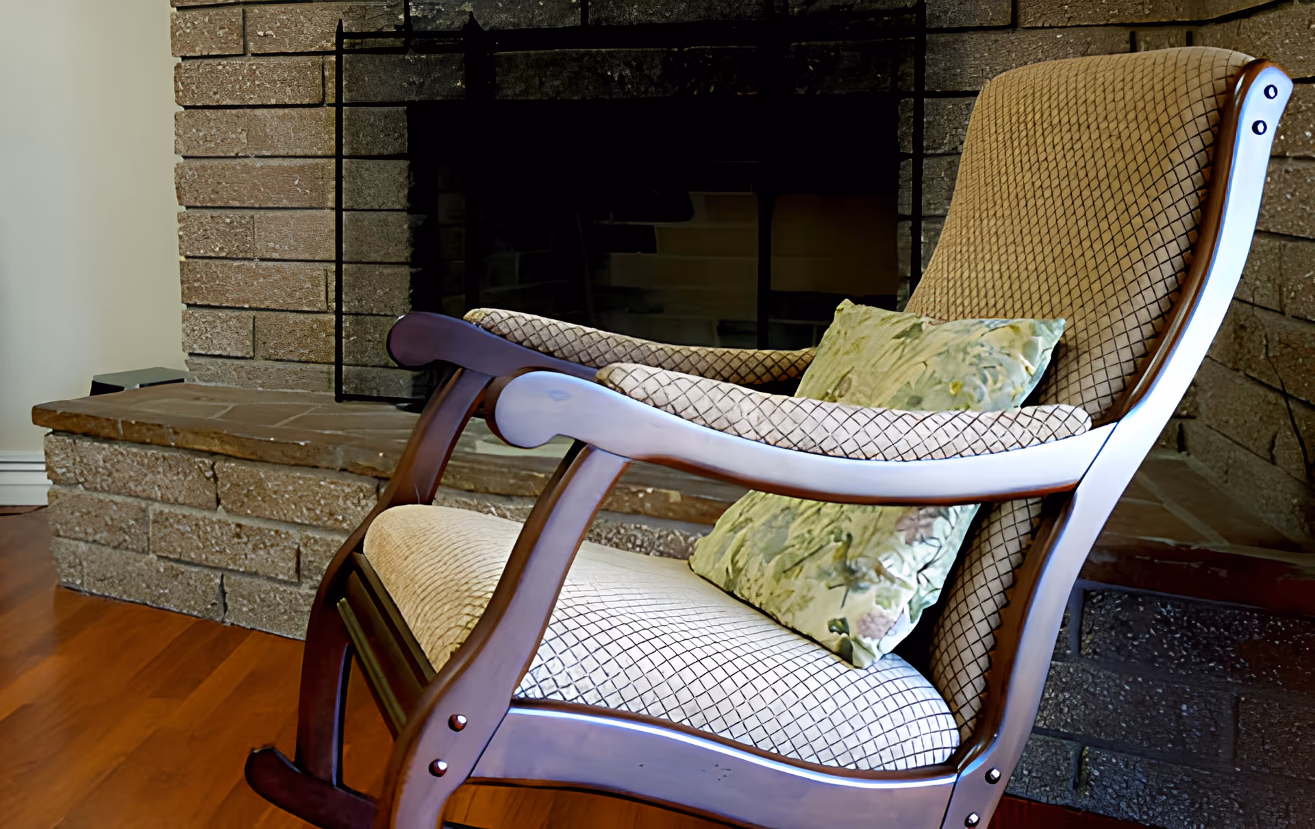 A cushioned wooden rocking chair with a patterned fabric and a floral pillow placed in front of a brick fireplace with a stone hearth in a room with wooden flooring.