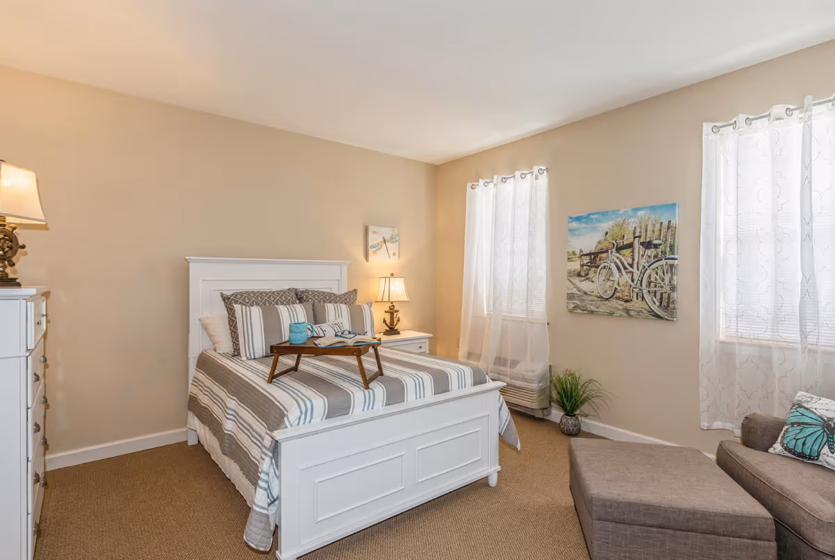 A cozy bedroom with a white wooden bed frame and striped bedding. There is a small wooden tray on the bed holding a blue mug and an open book. To the left of the bed is a white dresser with a lamp on top. On the right side, there is a nightstand with a lamp and a small decorative item. The room has beige walls, two windows with sheer white curtains, a painting of a bicycle on the wall, a small green plant near the corner, and a brown upholstered chair with a matching ottoman.