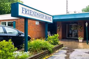Exterior view of a single-story brick building with a blue roof and a sign in front that reads 'FRIENDSHIP HOME'. There is a black car parked to the left, some green bushes and plants around the sign, and a wet pavement indicating recent rain.