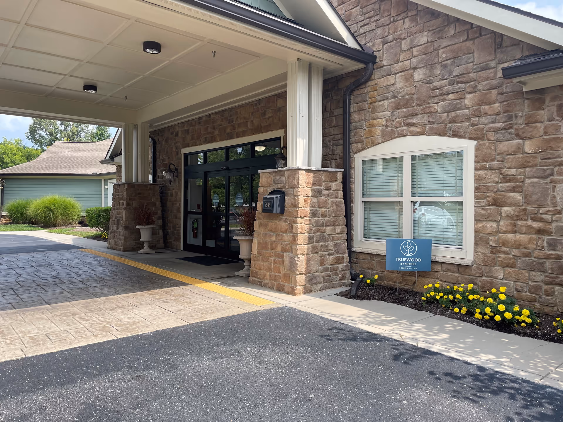 Entrance to a senior living facility with stone exterior walls, a covered driveway, and a glass door entrance. There are two large stone pillars supporting the roof over the driveway, potted plants on either side of the entrance, and a small garden bed with yellow flowers and a sign that reads 'TRUEWOOD BY MORRILL SENIOR LIVING'.