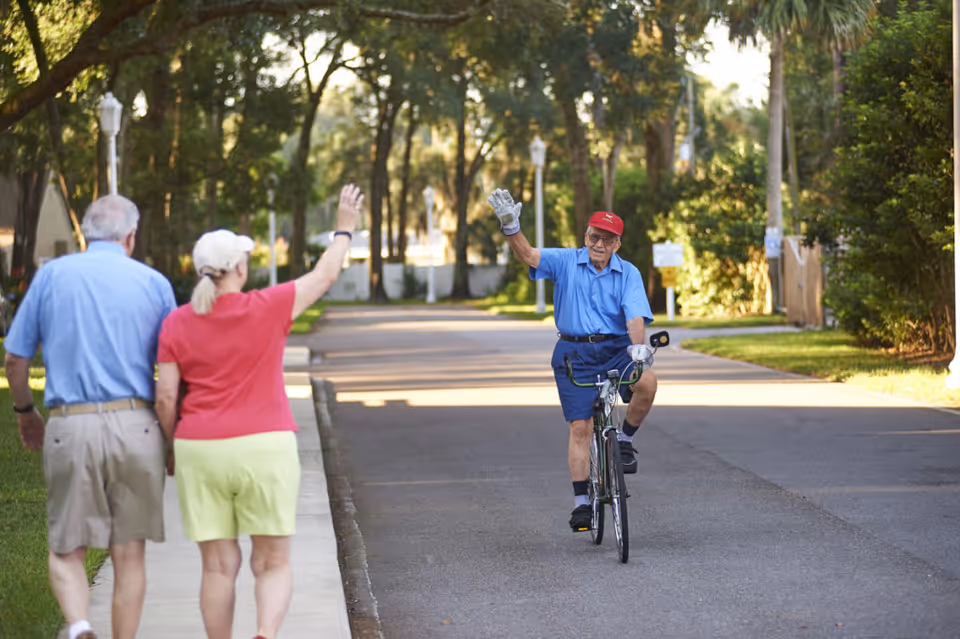 An elderly man wearing a red cap and blue shirt rides a bicycle on a paved road in a tree-lined neighborhood, waving with his right hand. Two other elderly individuals, a man and a woman, walk on the sidewalk beside the road, with the woman waving back. The scene is bright and sunny with greenery and street lamps visible.