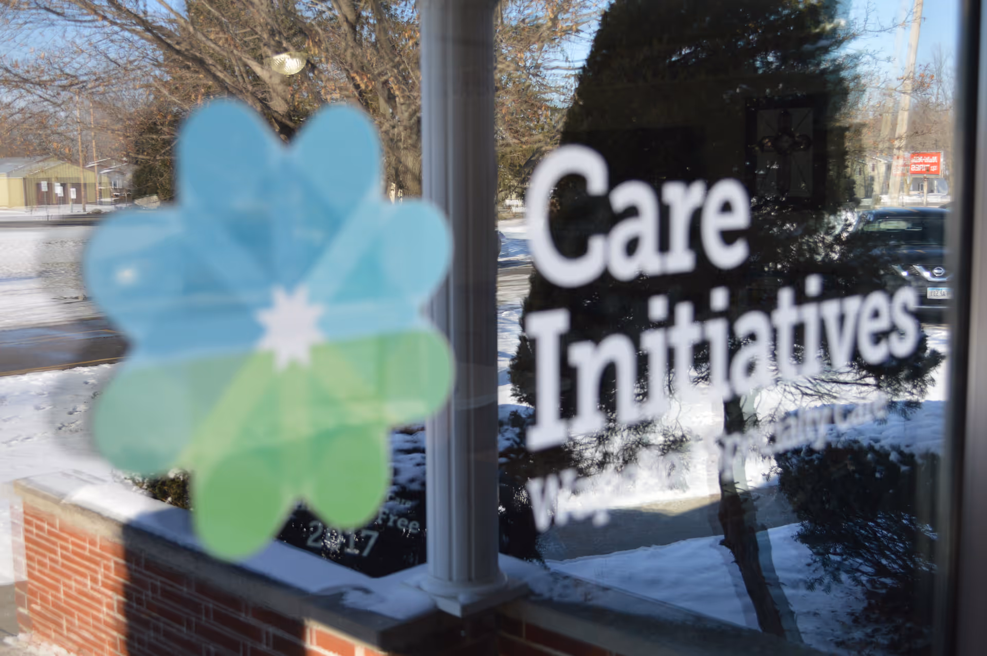 A glass window with a colorful flower-shaped logo and the words 'Care Initiatives' printed on it. Outside, there is a snowy landscape with trees, a brick ledge, and a street with parked cars.
