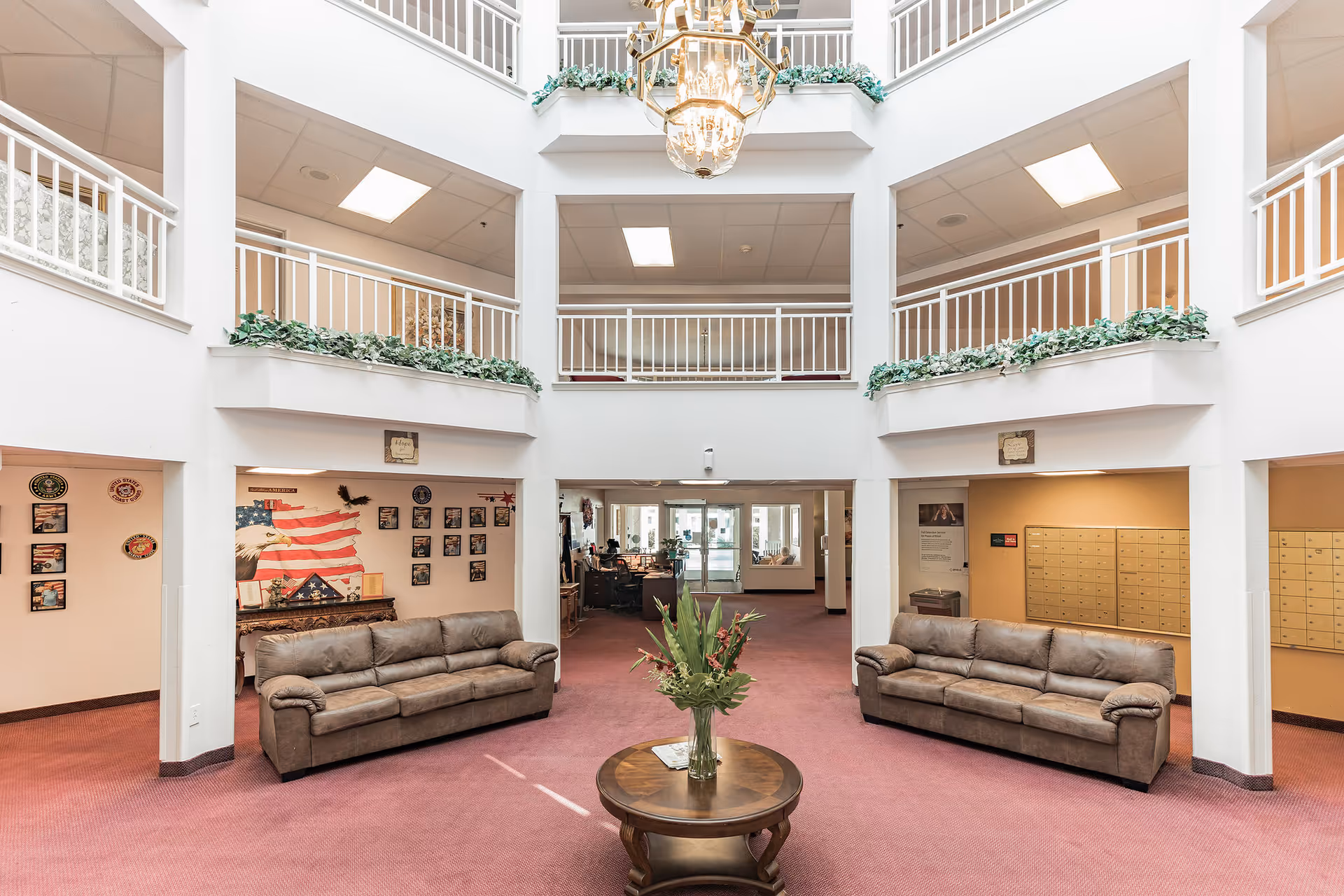 Spacious senior living community lobby with two brown leather sofas facing each other on a red carpet. A round wooden table with a vase of flowers is centered between the sofas. The lobby has white walls, a high ceiling with a large chandelier, and upper-level balconies with white railings and green plants. Mailboxes are visible on the right side, and a patriotic wall display with an American flag and eagle is on the left.