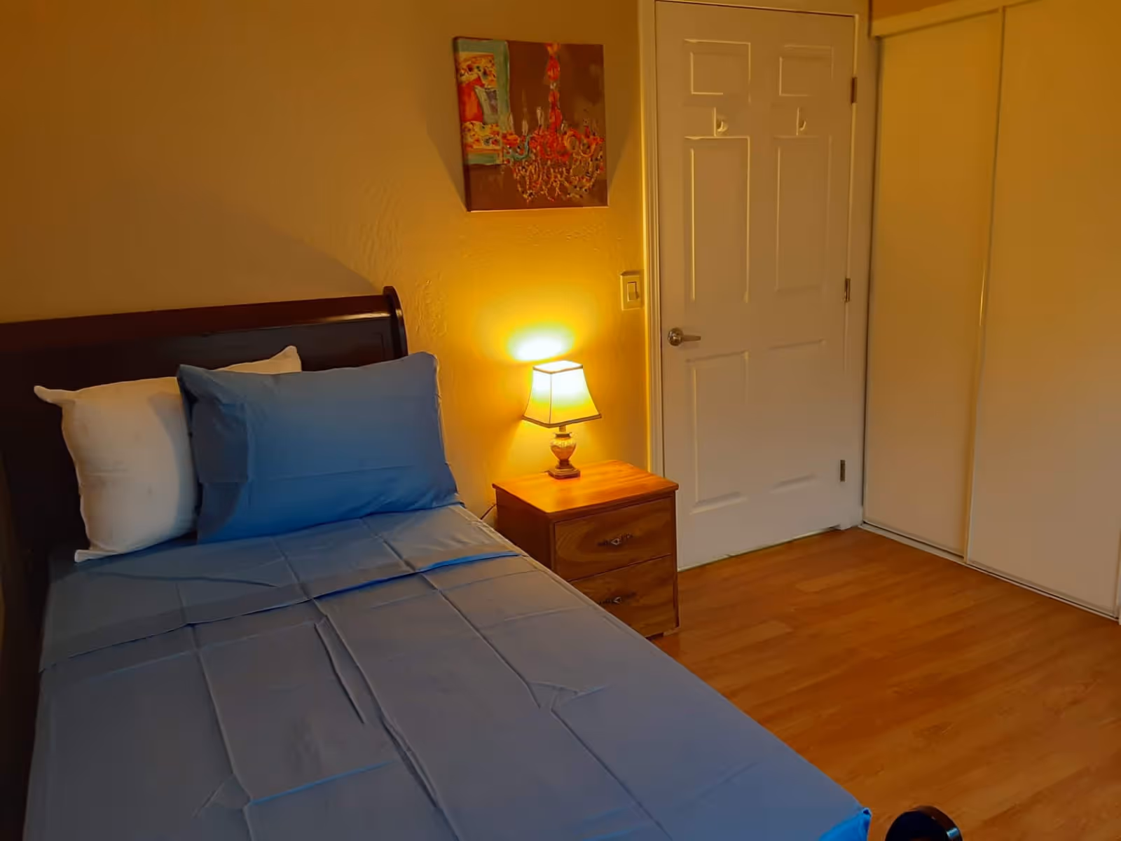 A tidy bedroom with a single bed dressed in blue sheets, a wooden nightstand with a lit lamp, and a closed door.