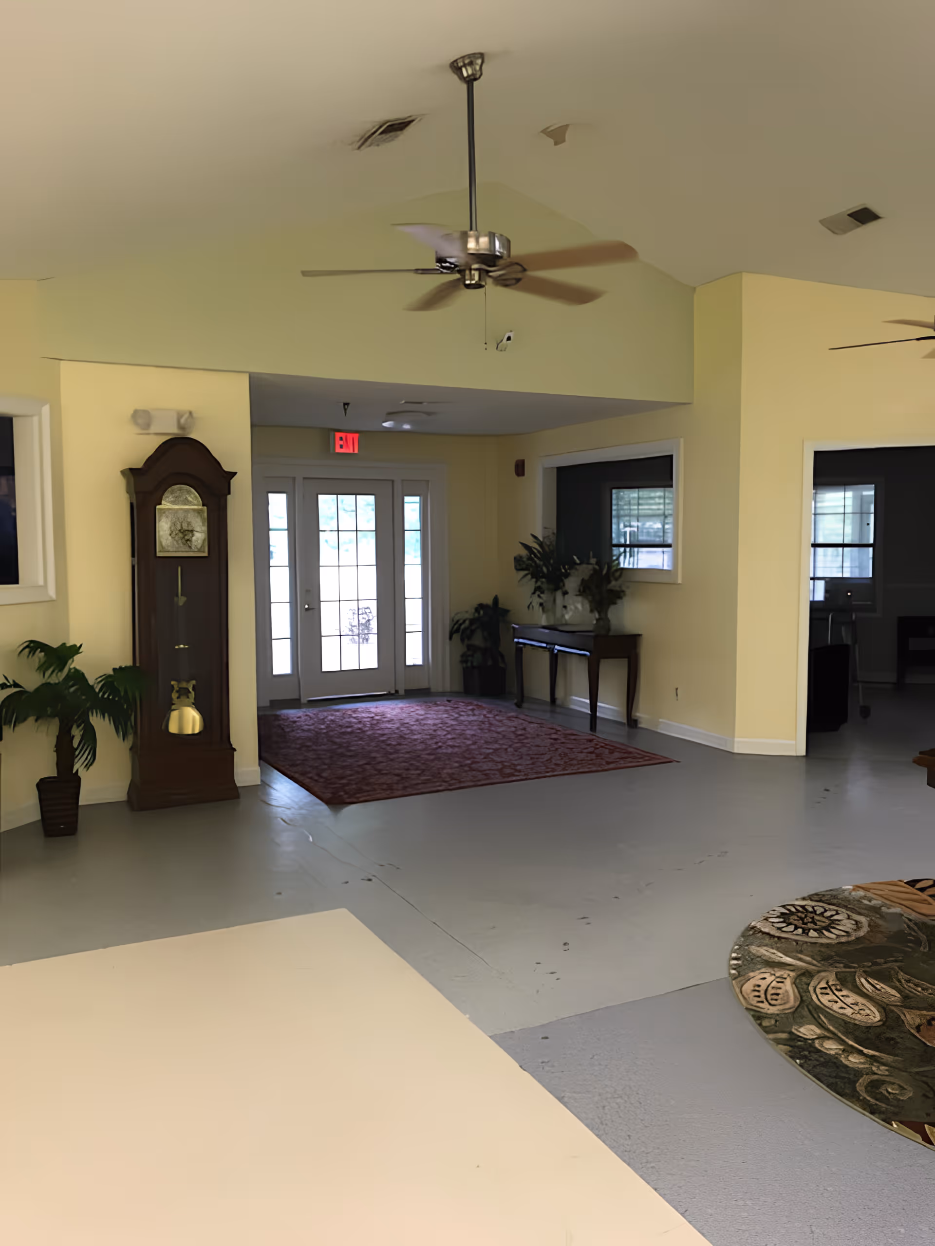 Entryway/common room interior with double glass doors, a grandfather clock, ceiling fans, plants and rugs.