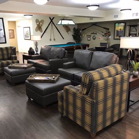 A cozy lounge area featuring comfortable seating with plaid and leather armchairs, a pool table in the background, and decorative wall art including fishing gear.