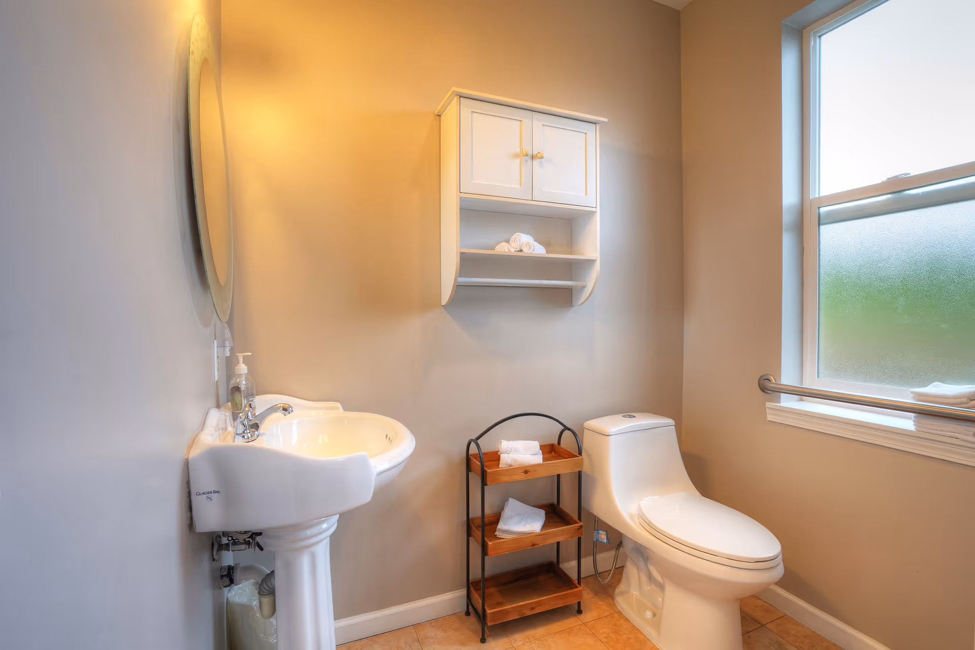 Bright bathroom with a pedestal sink and round mirror, wall cabinet and small towel shelf beside a toilet under a frosted window with a grab bar.