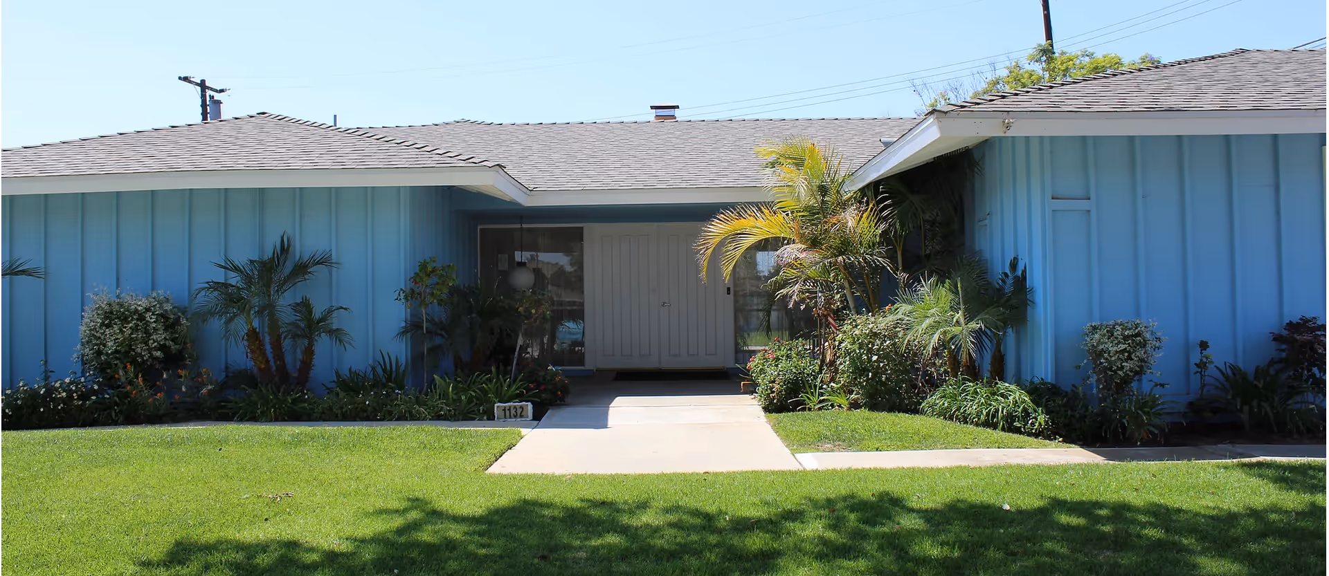 Front entrance of a single-story blue building with a concrete walkway, green lawn, and tropical landscaping.