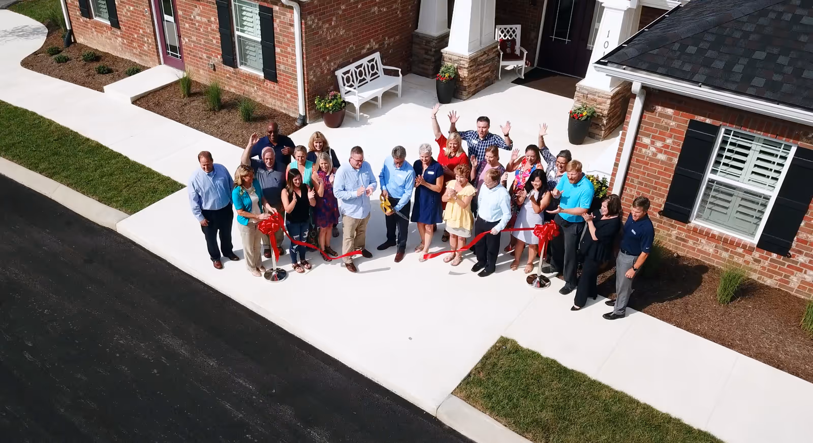 A group of people gathered outside the entrance of a brick building, participating in a ribbon-cutting ceremony. The group is standing on a white concrete walkway with some greenery and potted plants near the building. Several people are waving and smiling at the camera.