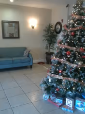 A decorated Christmas tree with lights, red and gold ornaments, and ribbon garland stands in a tiled room. Several wrapped presents are placed under the tree. To the left, there is a blue sofa with a patterned pillow, a framed picture on the wall, a wall-mounted light fixture, and a potted plant in the corner.