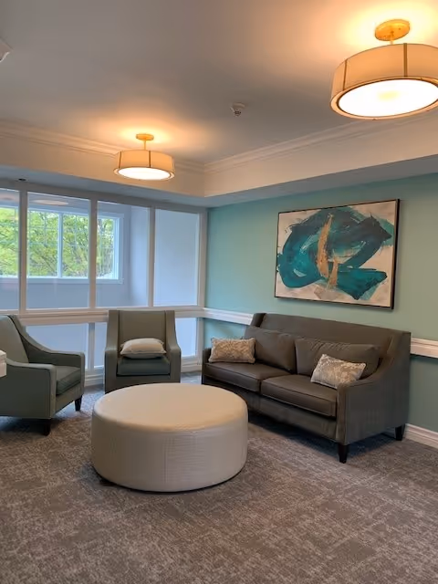 A cozy living room with a gray sofa, two gray armchairs, and a round beige ottoman in the center. The walls are painted light blue with white trim, and a large abstract painting with teal and gold colors hangs above the sofa. Two ceiling lights with round beige lampshades illuminate the room. Large windows on one side let in natural light and show greenery outside.