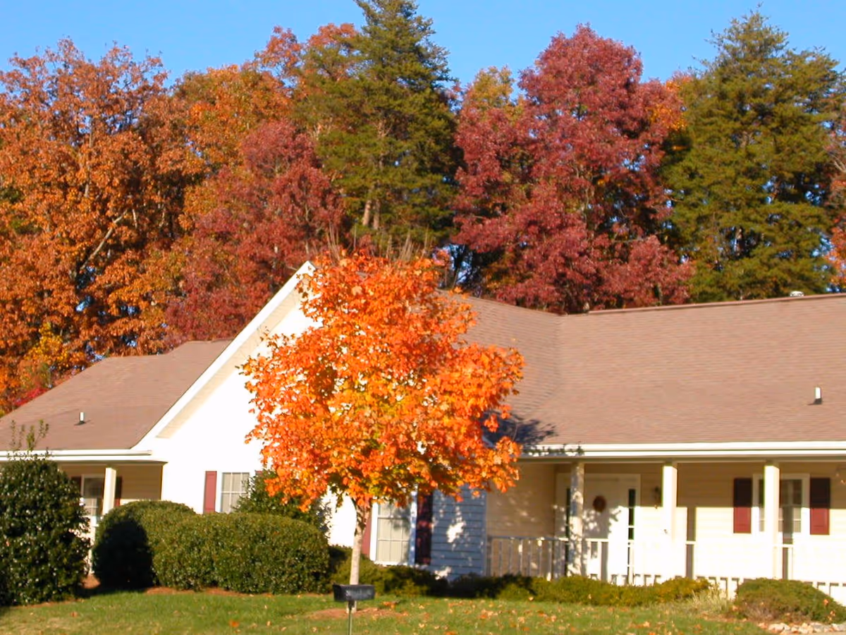 Front of a single-story residential building with a porch and a bright orange autumn tree on the lawn, set against colorful fall foliage.