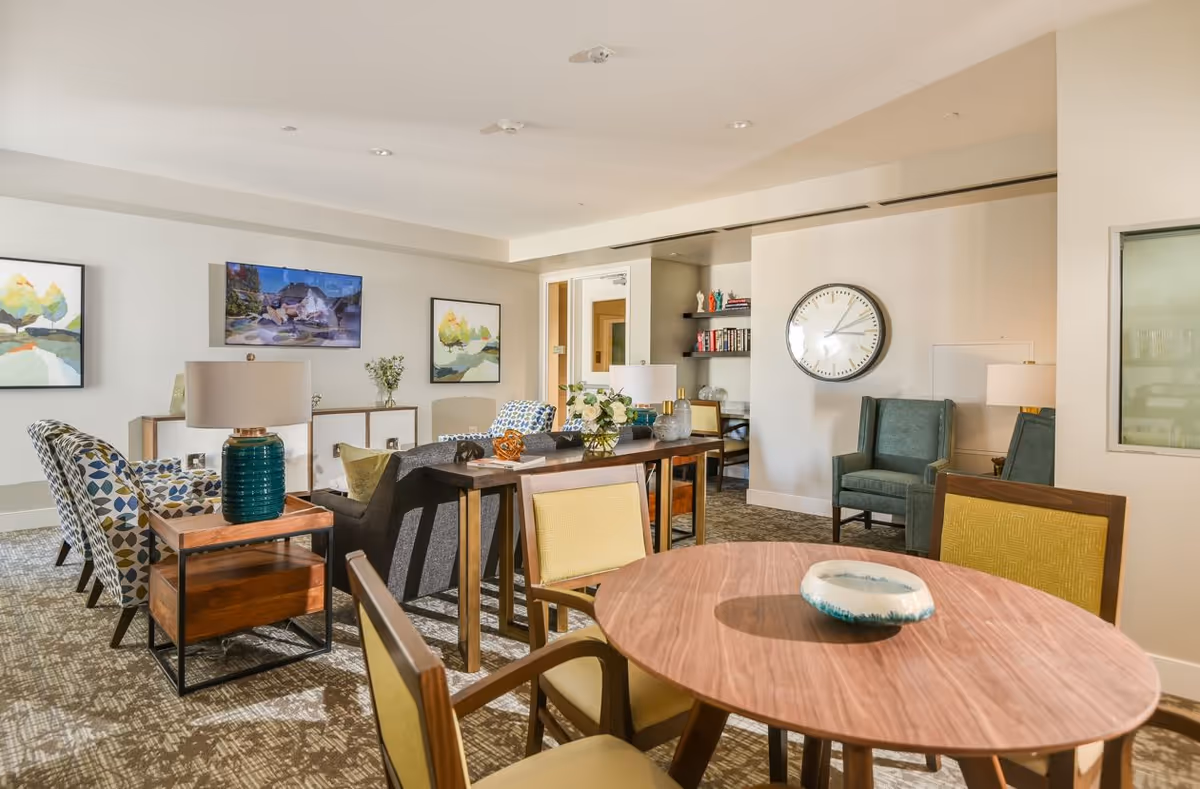 A bright and spacious senior living common area with a round wooden table and four chairs in the foreground. Behind the table is a seating area with patterned armchairs, a gray sofa, side tables with lamps, and decorative items. The walls are adorned with framed artwork and a large clock. The room has a carpeted floor and a neutral color palette with pops of color from the furniture and decor.