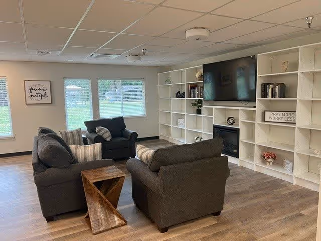 A cozy living room area in a senior community with three dark gray upholstered chairs arranged around a wooden side table. The room features large windows with blinds, a white built-in bookshelf filled with books and decorative items, a flat-screen TV mounted above a fireplace, and light wood flooring.