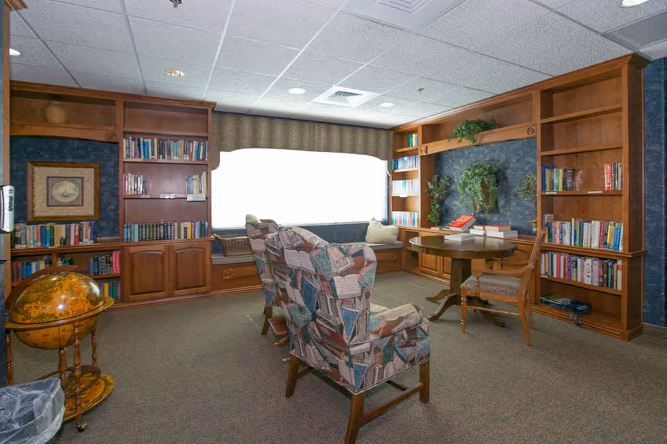A cozy reading room with wooden bookshelves filled with books, two patterned armchairs, a round wooden table with chairs, a globe on a stand, and a large window with a cushioned bench underneath. The room has carpeted floors and a ceiling with recessed lighting.