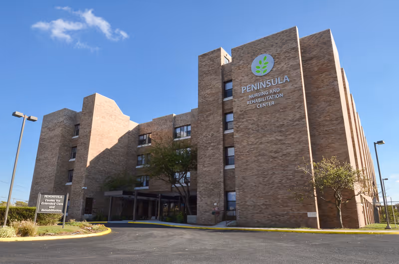 Exterior view of a multi-story brick building with the sign 'PENINSULA Nursing and Rehabilitation Center' on the front. There is a driveway and some landscaping with trees and bushes in front of the building under a clear blue sky.