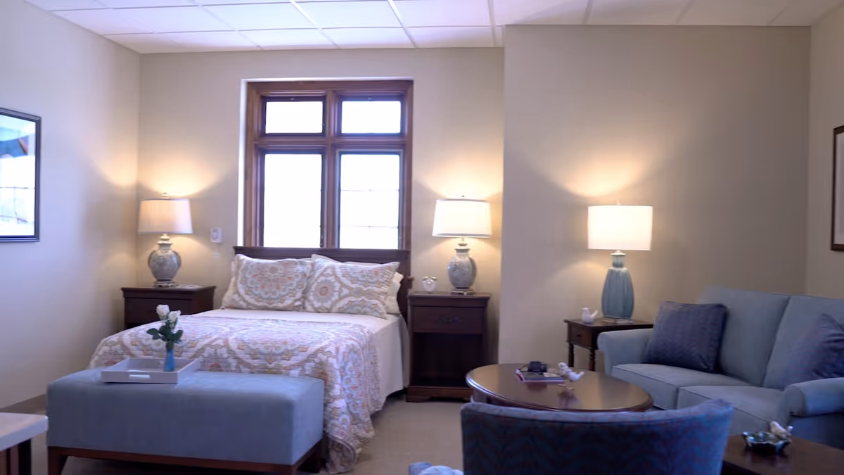 Bright bedroom suite with a double bed flanked by nightstands and lamps, plus a sofa, coffee table, and ottoman.
